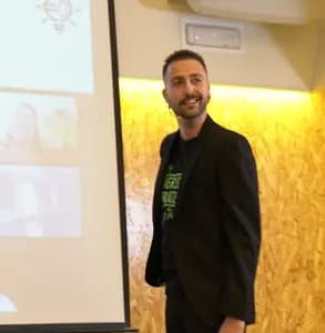 A smiling man with short dark hair and a beard stands in front of a projection screen displaying a slide with a lightbulb icon and blurred images, wearing a black blazer over a green graphic t-shirt, with a warmly lit textured wooden wall behind him, suggesting a presentation or educational setting.